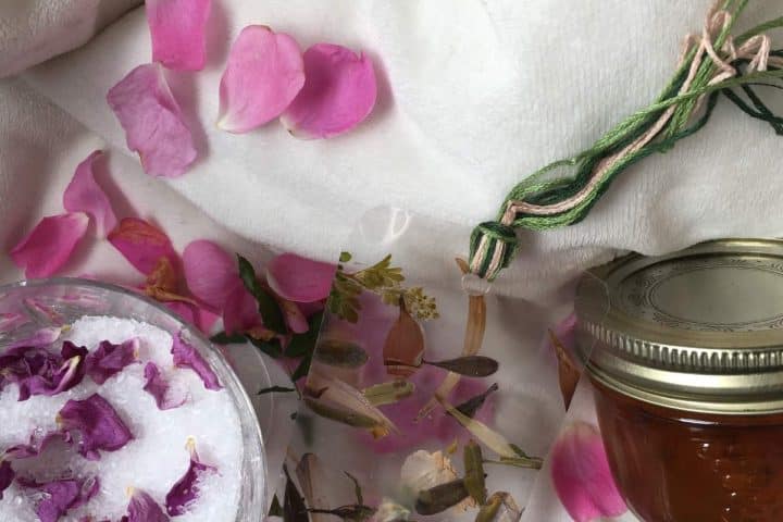A close-up of a flatlay composition featuring rose petals, a bowl of bath salts with flowers, dried herbs, a green and pink braided cord, and a jar with a metal lid on a soft fabric background.