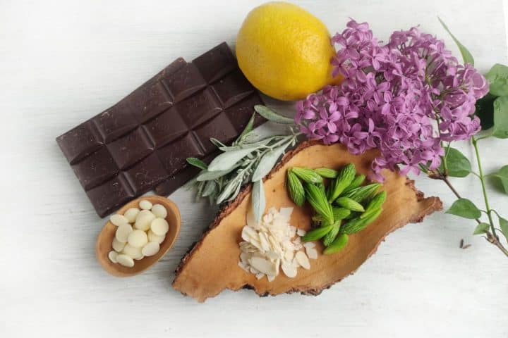 A bar of dark chocolate, a lemon, purple flowers, sage leaves, white chocolate buttons in a bowl, sliced almonds, and green plant buds on a wooden surface.
