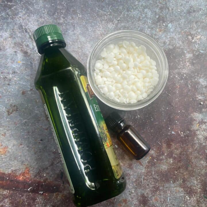 A bottle of olive oil next to a bottle of white beeswax pellets and a small bottle of essential oils.