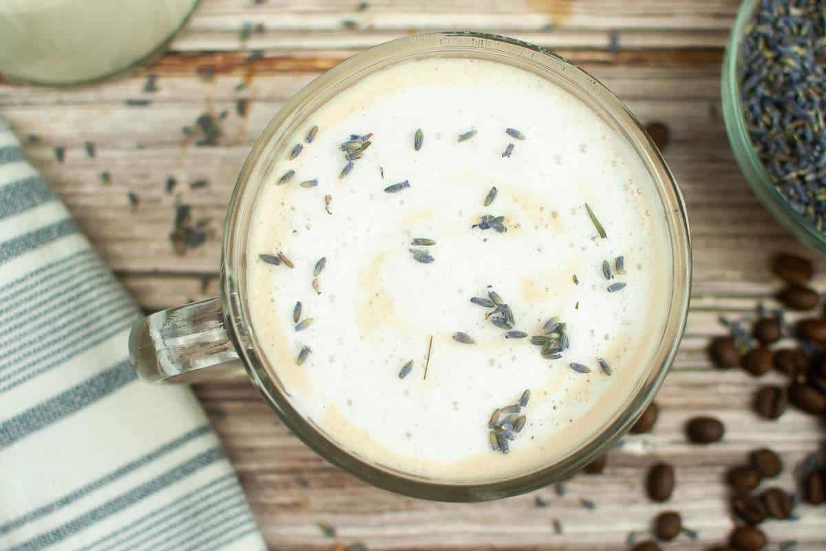 A top-down view of a mug filled with a frothy, lavender-infused beverage, surrounded by scattered coffee beans and lavender buds on a wooden surface.
