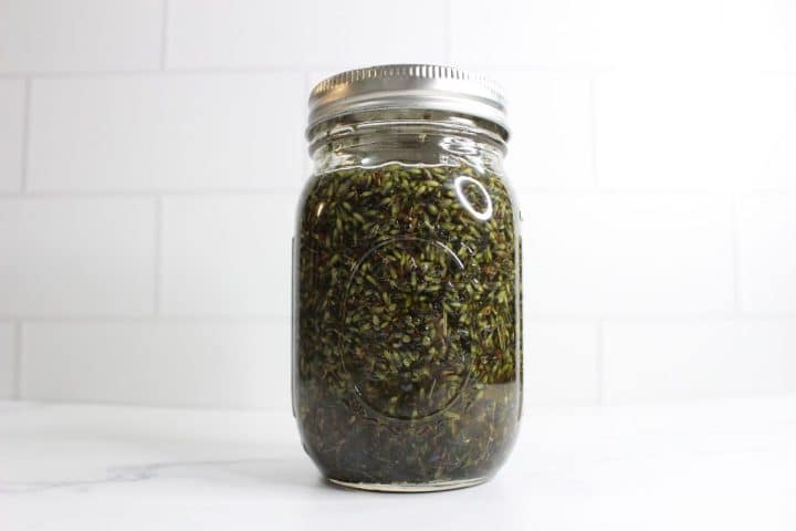 A glass jar filled with a green leafy substance, sealed with a metal lid, placed on a white surface in front of a white tiled wall.