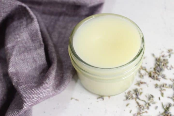 A small glass jar filled with creamy balm next to a folded purple cloth and scattered dried herbs on a white surface.
