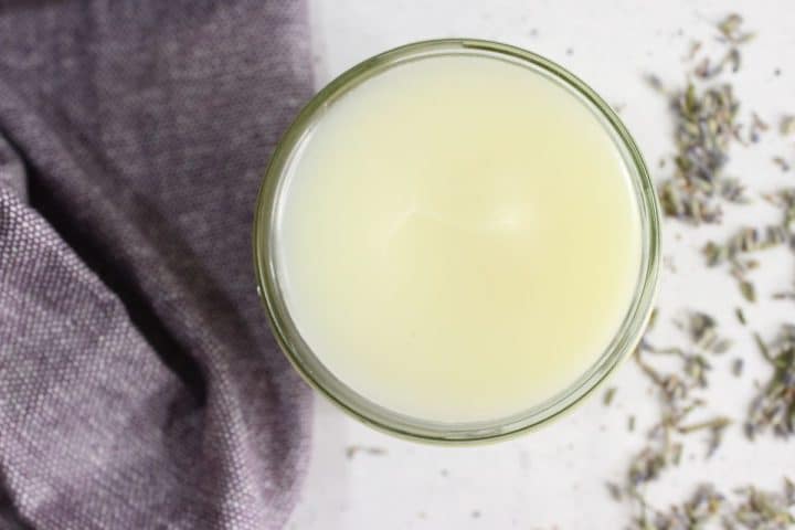 Top view of a glass filled with a light yellow liquid on a white surface, next to a purple fabric and scattered dried lavender flowers.