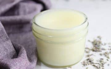 A small glass jar filled with lavender tallow balm sits on a surface with scattered lavender buds and a purple cloth nearby.