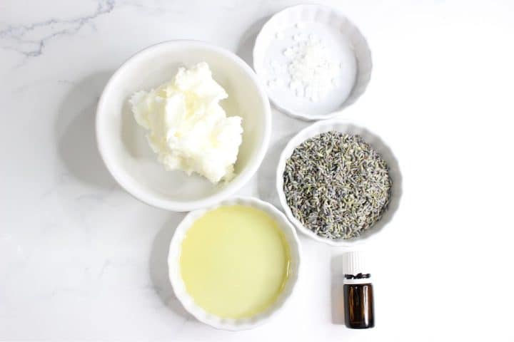 Five bowls contain shea butter, small white granules, dried lavender, oil, and a small bottle on a marble surface.