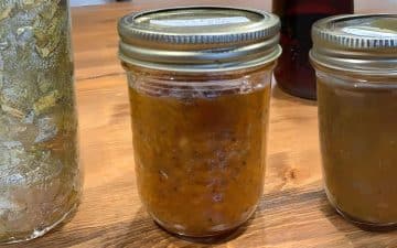 Three jars of homemade preserves on a wooden table, with one showing chunky orange marmalade and others appearing less textured.