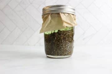 Glass jar filled with dried herbs, covered with paper and sealed with a metal lid, placed on a white surface with a tiled background.