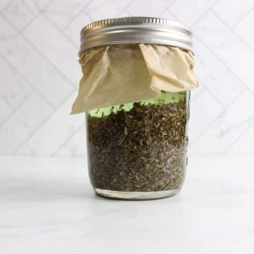 Glass jar filled with dried herbs, covered with paper and sealed with a metal lid, placed on a white surface with a tiled background.
