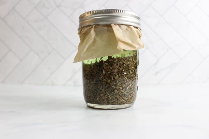 Glass jar filled with dried herbs, covered with paper and sealed with a metal lid, placed on a white surface with a tiled background.