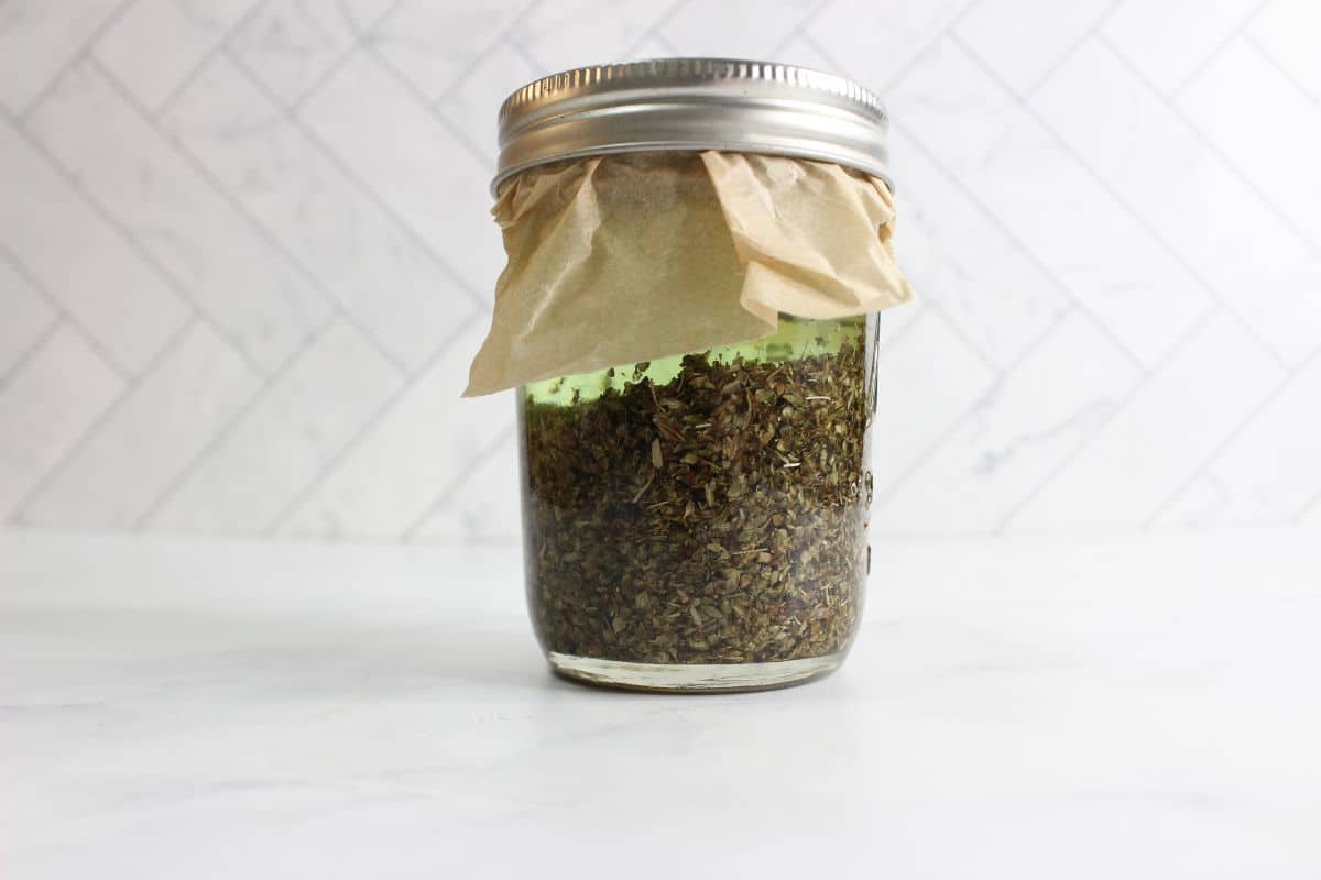 Glass jar filled with dried herbs, covered with paper and sealed with a metal lid, placed on a white surface with a tiled background.
