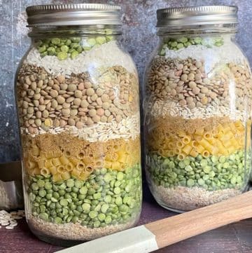 Two jars filled with layers of various dried ingredients, including lentils, pasta, and peas, with a measuring cup and wooden spatula beside them. Mason jar soup mix recipe.