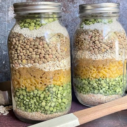 Two jars filled with layers of various dried ingredients, including lentils, pasta, and peas, with a measuring cup and wooden spatula beside them. Mason jar soup mix recipe.