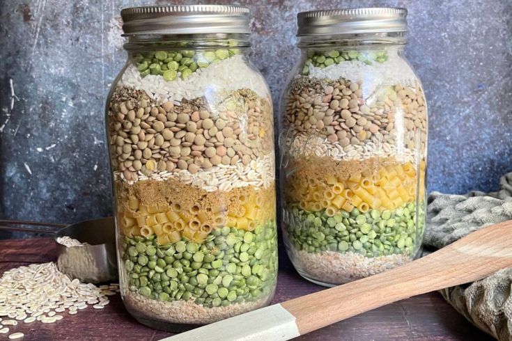 Two jars filled with layers of various dried ingredients, including lentils, pasta, and peas, with a measuring cup and wooden spatula beside them. Mason jar soup mix recipe.