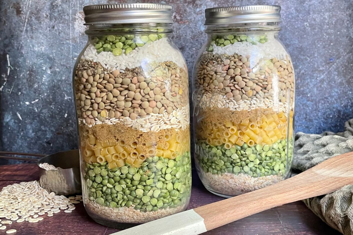 Two jars filled with layers of various dried ingredients, including lentils, pasta, and peas, with a measuring cup and wooden spatula beside them. Mason jar soup mix recipe.