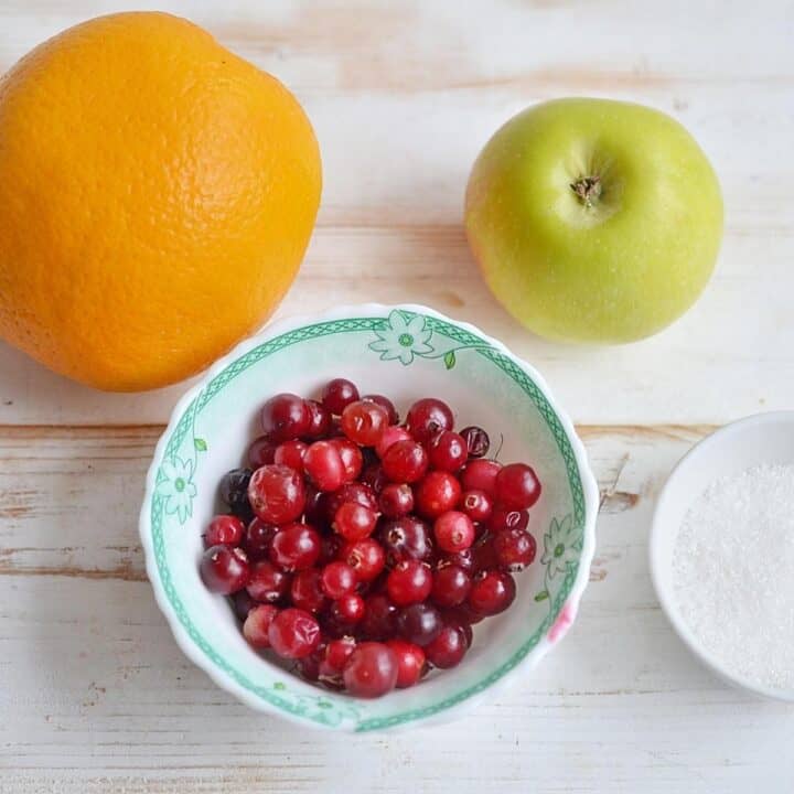 No Cook Cranberry Relish Recipe ingredients - orange, cranberries, apple, and sugar on a wooden background.