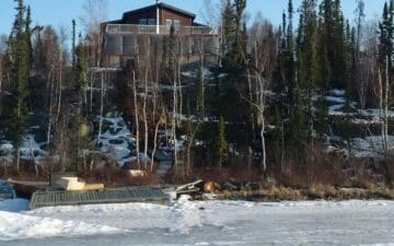 An off grid house sits on top of an ice covered hill down to a lake with an exposed water pipe.