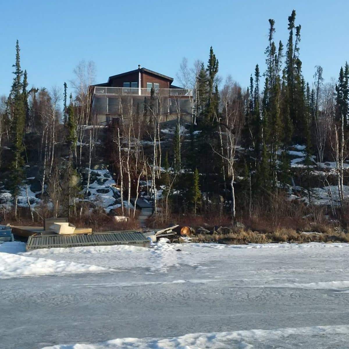 An off grid house sits on top of an ice covered hill down to a lake with an exposed water pipe.