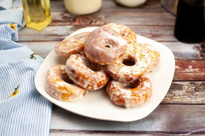 A plate of glazed donuts stacked on a wooden table with a striped cloth and bottles nearby.
