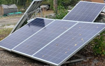 Three solar panels installed on metal frames are positioned at an angle on the ground in an outdoor setting surrounded by vegetation and rocks.