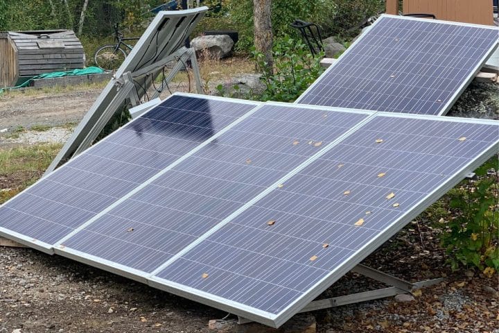 Three solar panels installed on metal frames are positioned at an angle on the ground in an outdoor setting surrounded by vegetation and rocks.