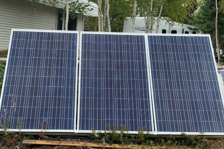 Three solar panels are mounted in front of a building and surrounded by vegetation.