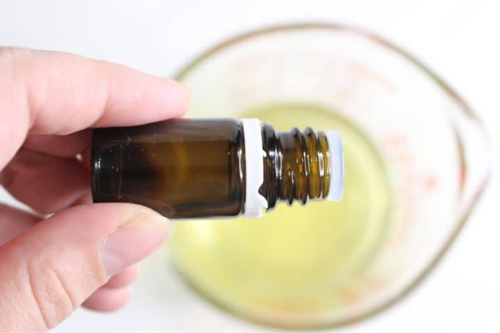 A hand holds a small amber glass bottle above a clear measuring cup filled with yellow liquid.