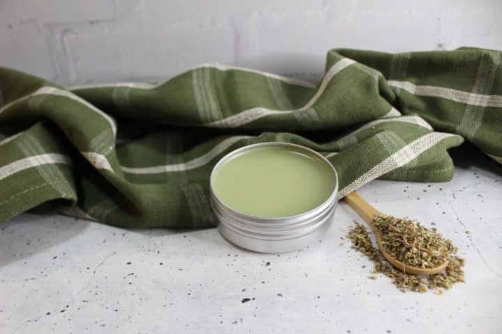 A tin of green balm sits on a speckled surface next to a wooden spoon with dried herbs. A green plaid cloth is draped in the background.