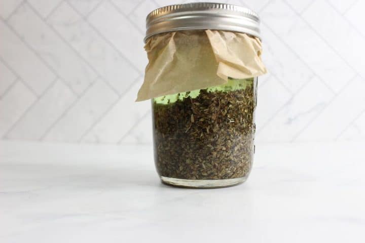 A jar filled with dried herbs, sealed with a paper cover and a metal lid, placed on a white marble surface with a subtle tiled background.