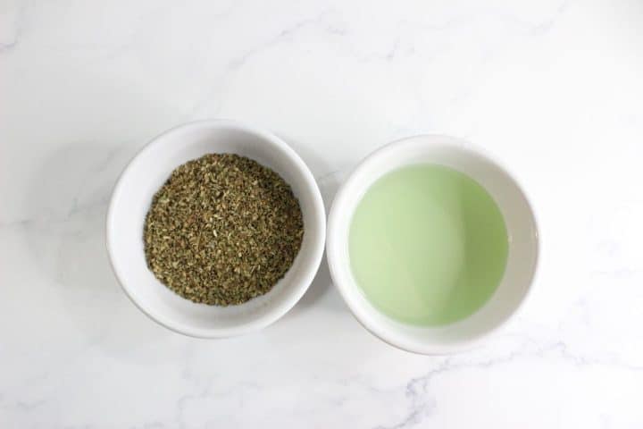Two white bowls on a marble surface; the left contains dried oregano, the right has a green liquid.