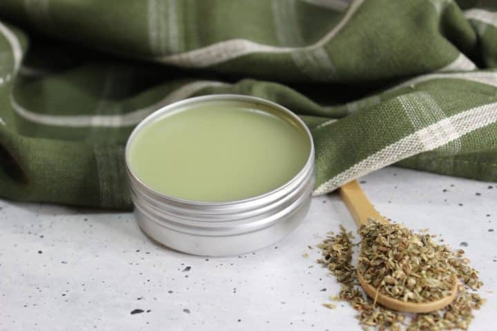 A small tin of green herbal balm is open on a speckled surface. A wooden spoon with dried herbs is beside it. A green cloth with white stripes is in the background.