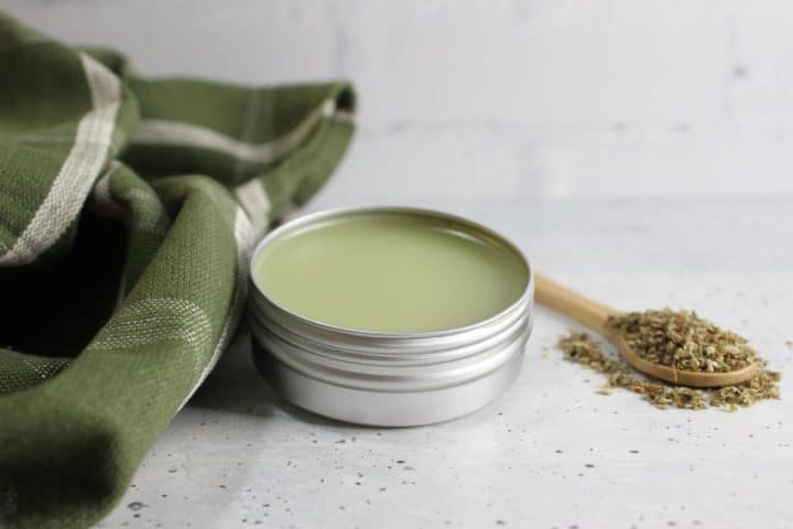 A metal tin filled with green salve is placed next to a wooden spoon containing dried herbs. A green cloth is positioned alongside on a light speckled surface.