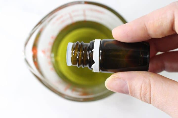 Hand holding a small brown bottle above a measuring cup filled with green liquid.