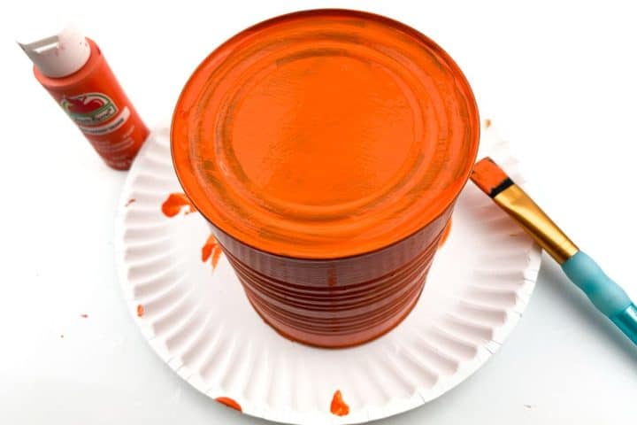 An orange-painted tin can on a paper plate, with a paintbrush and orange paint bottle nearby.