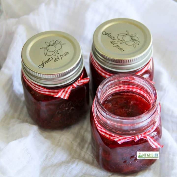 Three glass jars filled with red jam sit on a white cloth; two have metal lids and all are decorated with red and white ribbon, perfect for trying your favorite strawberry syrup recipe.