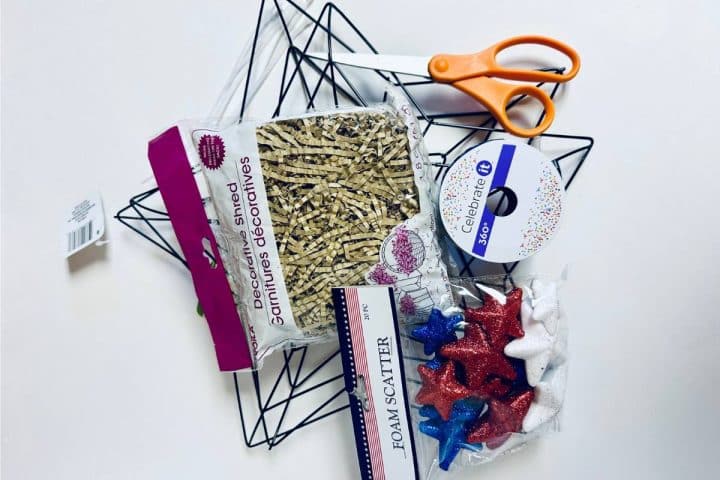 A collection of crafting supplies including shredded paper, decorative foam stars, ribbon, scissors, and a wire frame on a white background.
