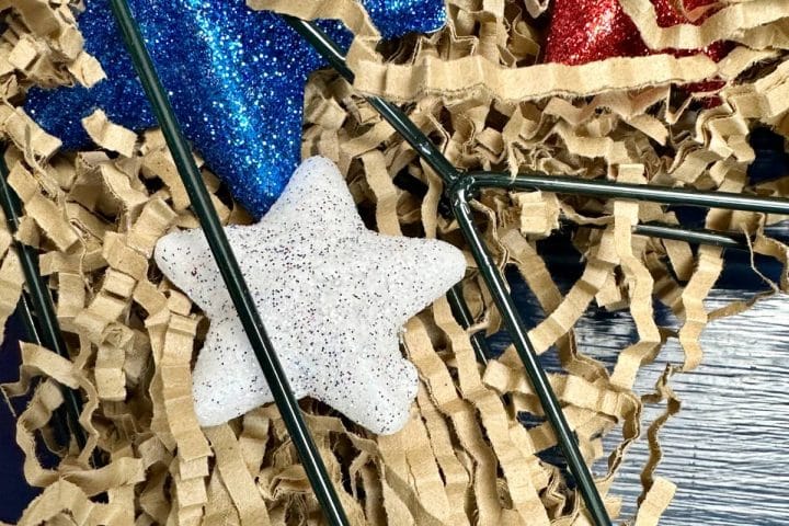 A white glittery star ornament is placed among red and blue glittery star ornaments and shredded brown paper packing material.