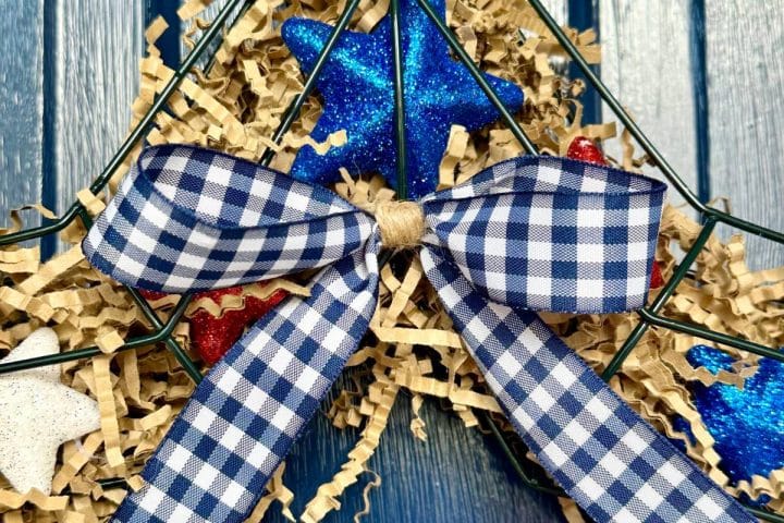 A close-up of a decorative wreath featuring a blue and white checkered bow, glittery blue and red stars, and shredded paper on a blue wooden background.