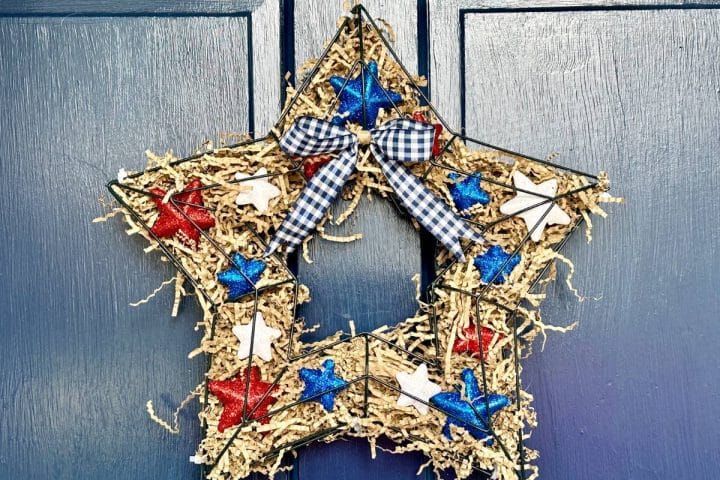 A rustic DIY patriotic wreath swith red, white, and blue accents, adorned with a black and white gingham ribbon, hanging on a blue door.