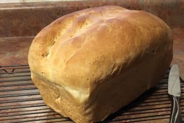 A loaf of freshly baked bread rests on a cooling rack next to a butter knife on a brown countertop.
