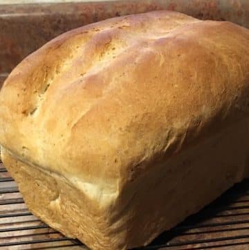 A loaf of freshly baked bread rests on a cooling rack next to a butter knife on a brown countertop.