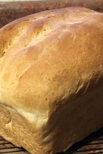 A loaf of freshly baked bread rests on a cooling rack next to a butter knife on a brown countertop.