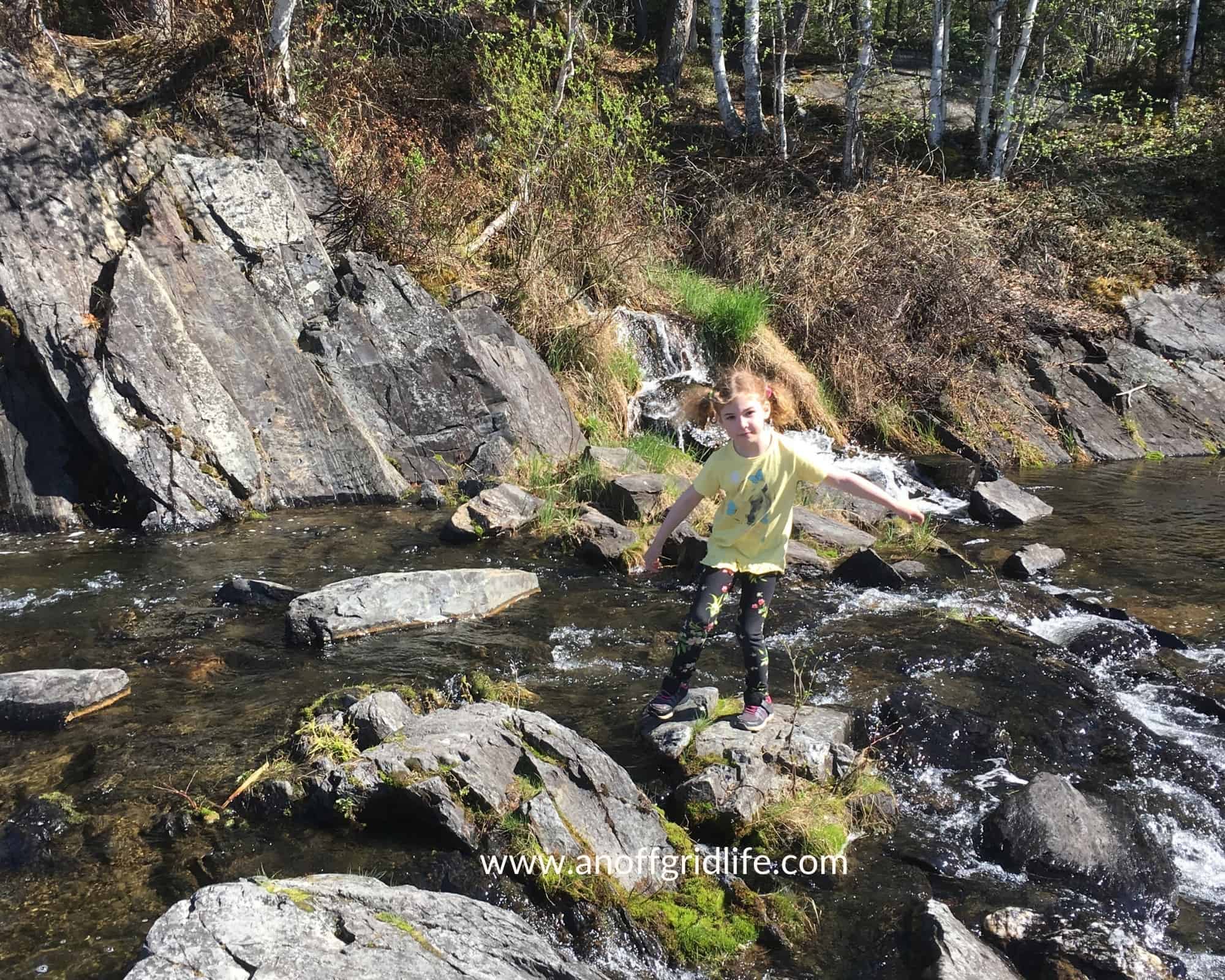A girl learning wilderness survival skills stands on rocks near a stream.