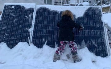 A little girl standing next to a snow-covered solar panel, showcasing the potential to survive off the grid with renewable energy.