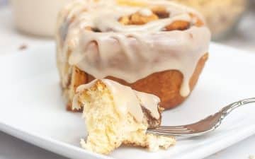 A close-up of a sourdough cinnamon roll with white icing on a white plate. A bite has been taken out, and a piece is on a fork in front.
