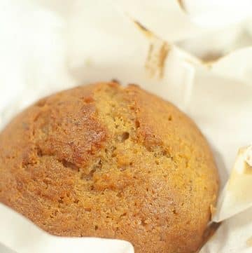 A freshly baked muffin wrapped in white parchment paper, showing a golden-brown top with a slightly cracked surface.