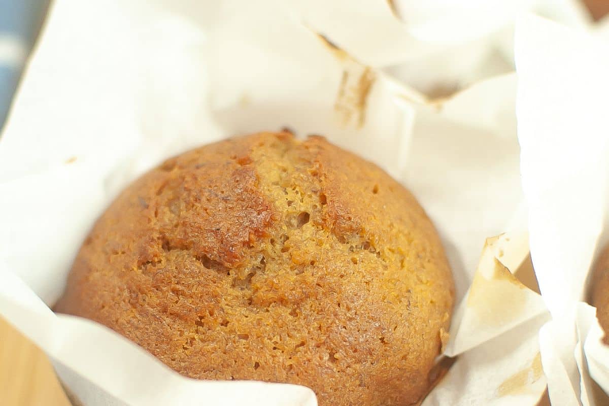 A freshly baked muffin wrapped in white parchment paper, showing a golden-brown top with a slightly cracked surface.