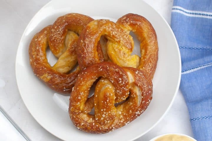 A white plate with three salted soft pretzels next to a blue cloth napkin.