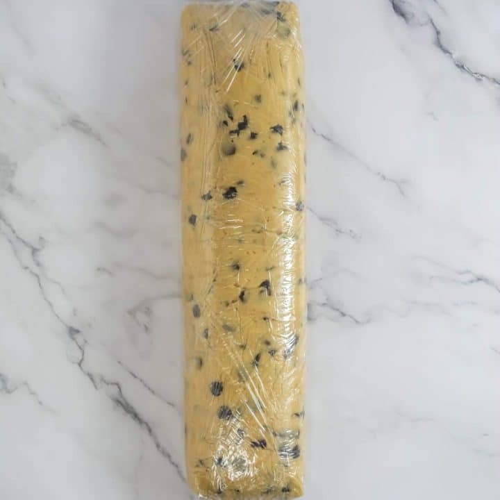 A plastic wrapped roll of sourdough discard chocolate chip cookies on a marble surface.
