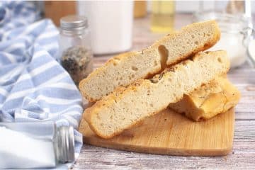 Two slices of sourdough focaccia rest on a wooden cutting board, accompanied by a blue striped cloth and an array of seasoning containers in the background.