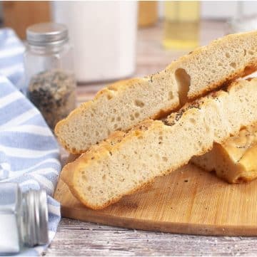 Two slices of sourdough focaccia rest on a wooden cutting board, accompanied by a blue striped cloth and an array of seasoning containers in the background.